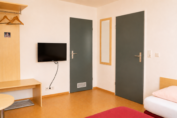 Ein einfaches Zimmer mit zwei Türen, Fernseher, Tisch und einem Bett.