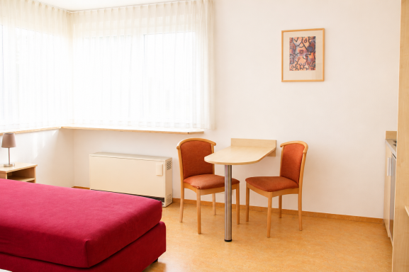 Gemütliches Zimmer mit rotem Bett, Tisch und zwei Stühlen, hellen Fenstern und Kunstwerk.