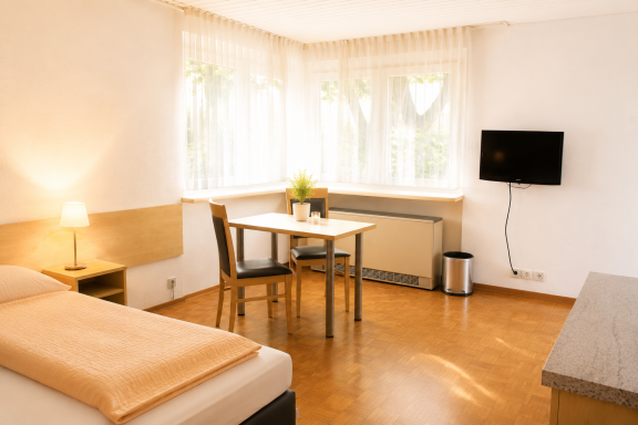 Helle Zimmer mit Bett, Tisch, Stühlen, TV und großen Fenstern.