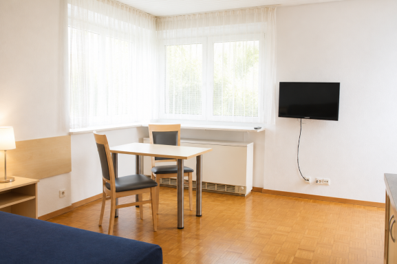 Heller Raum mit Tisch, Stühlen, Sofa und Wand-TV bei großen Fenstern.