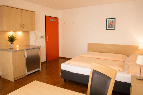 Ein einfaches Zimmer mit einem Bett, Schreibtisch, Kühlschrank und einem orangefarbenen Tür.