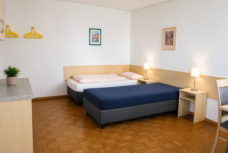 Helles Zimmer mit zwei Betten, Nachttischen und einem Tisch. Lampe und Pflanze vorhanden.